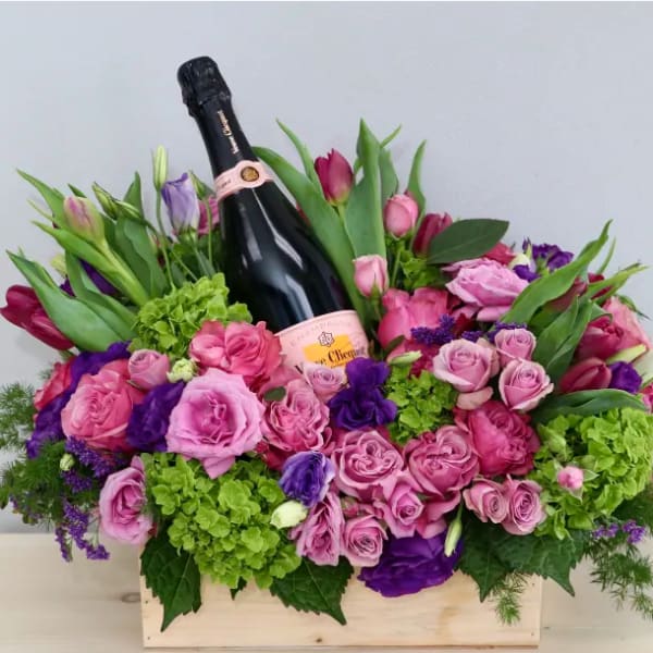 Wooden box arrangement of pink and purple flowers with a champagne bottle nestled in the center