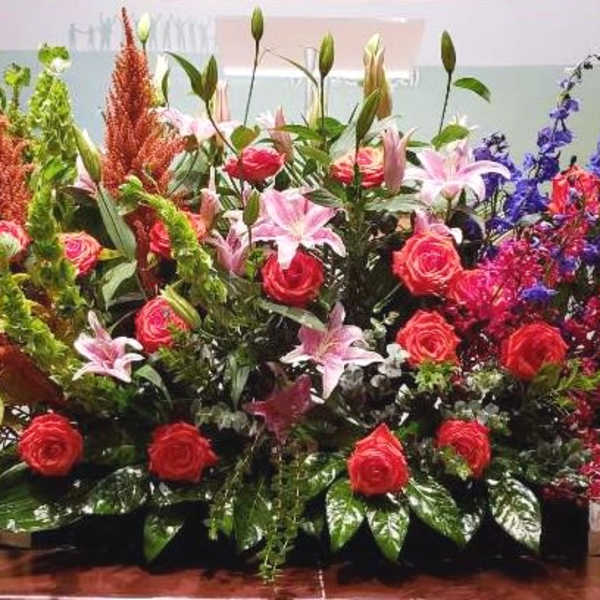 Large elongated arrangement of red roses, pink lilies, blue delphinium, and orange plumes in a low container