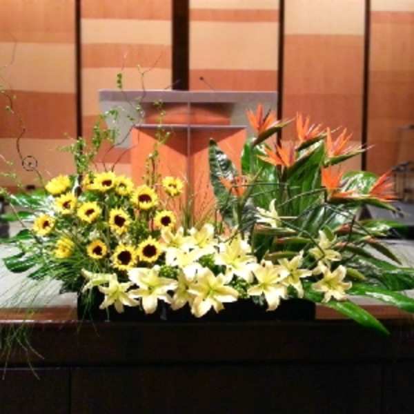 Long stage arrangement of yellow lilies, sunflowers, and orange tropical flowers in a low container
