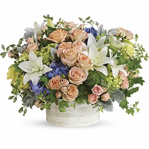 Low pastel arrangement of peach roses, white lilies, and blue hydrangeas in a round white container