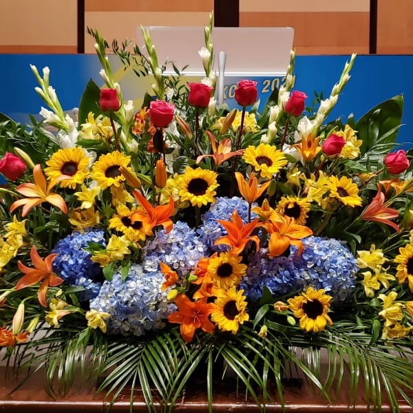 Large stage arrangement with yellow sunflowers, blue hydrangeas, orange lilies, and pink roses
