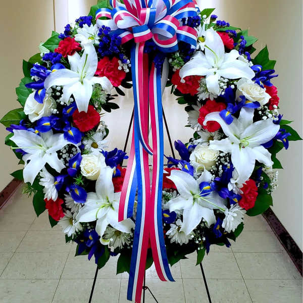 Large floral wreath with white lilies, red carnations, blue flowers, and a tricolor ribbon bow