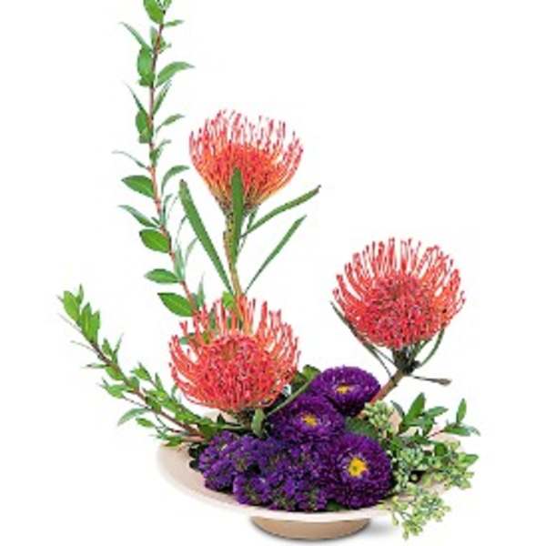 Modern arrangement of orange pincushion protea and purple flowers in a low ivory dish