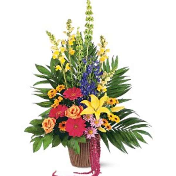 Tall mixed arrangement of yellow lilies, pink gerberas, roses, and blue delphinium in a brown pot with palm leaves.