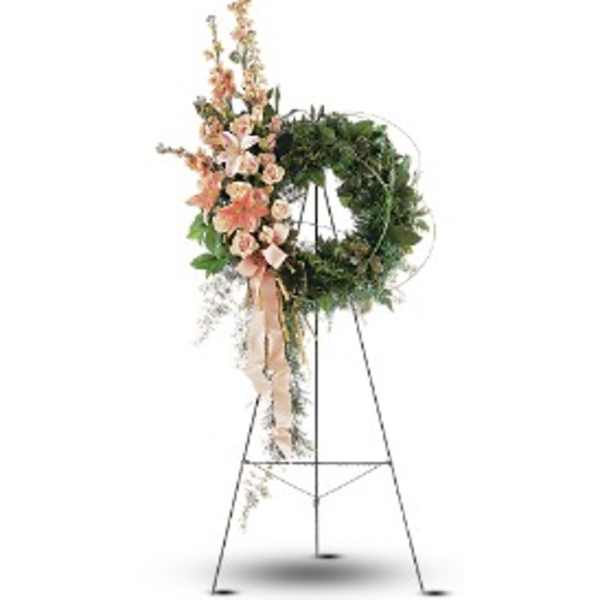Standing green wreath with peach lilies and carnations on a tall metal easel with cascading ribbon.