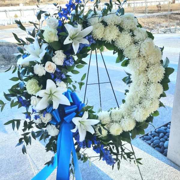 Standing white and blue funeral wreath with lilies and ribbon on a metal easel.