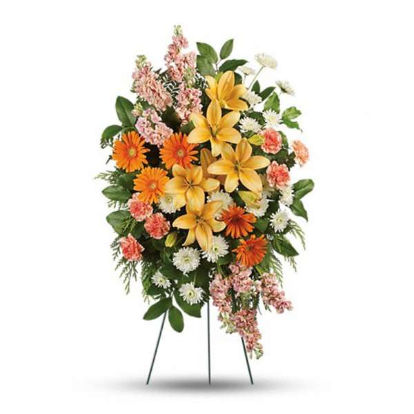 Oval standing spray of yellow lilies, orange gerberas, white mums, and pink flowers on a metal easel.