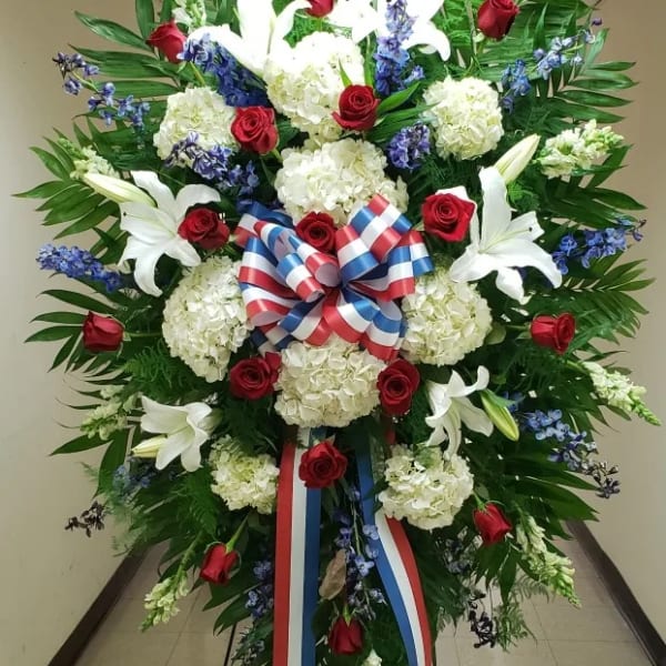 Tall standing spray of red roses, white lilies, white hydrangeas, and blue flowers with a red, white, and blue bow.