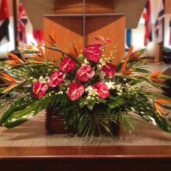 Large tropical arrangement with red blooms and orange accents in a low wooden container on a stage.