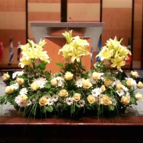 Low yellow lily, rose, and white daisy arrangement in a long container in front of a podium