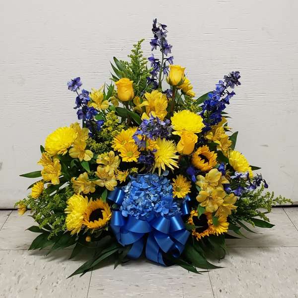 Fan-shaped yellow and blue arrangement with sunflowers, roses, delphinium, and a blue ribbon bow.