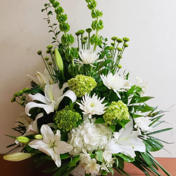 White and green floral arrangement with lilies, hydrangeas, mums, and bells of Ireland in a low white basket