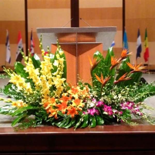 Large tropical stage arrangement with yellow spikes, orange lilies, and birds of paradise before a podium