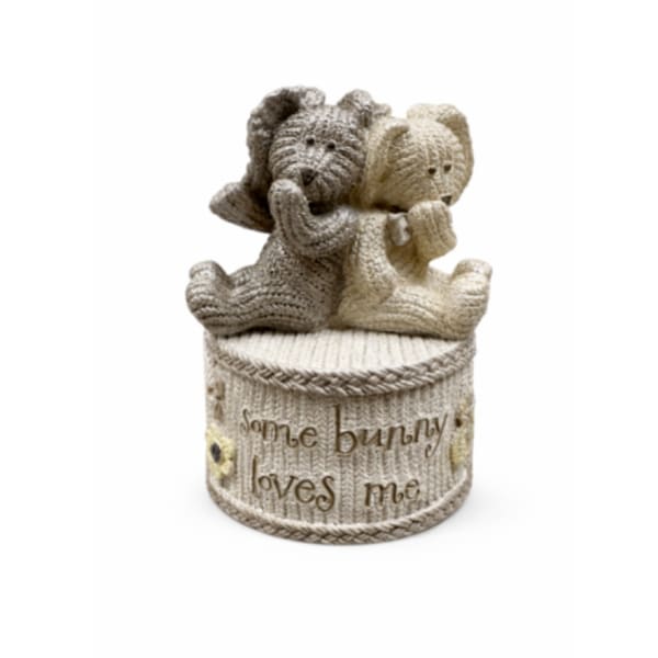 Two knitted teddy bears sit on a decorative container with text.