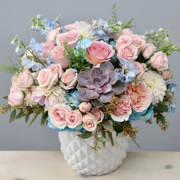 Pastel arrangement of pink roses, blue flowers, and a central succulent in a white ceramic vase
