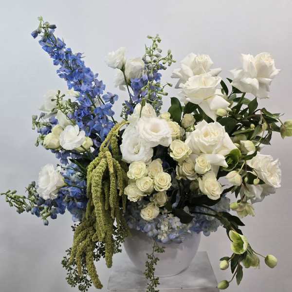 Tall white and blue flower arrangement with roses, lisianthus, and delphinium in a white bowl.