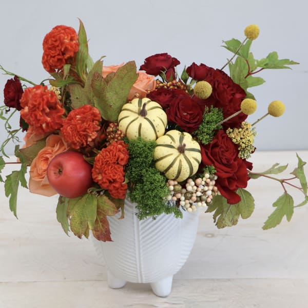 Autumn arrangement with red and orange flowers, mini pumpkins, and an apple in a white ceramic vase