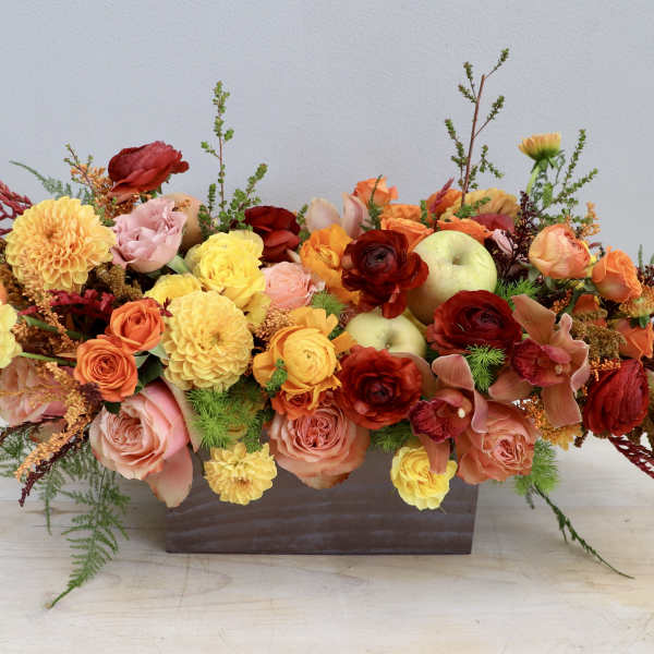 Low rectangular arrangement of orange, yellow, and red flowers with apples in a metal container