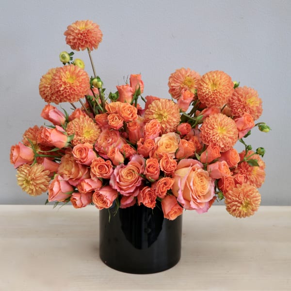 Low orange and peach rose and dahlia arrangement in a black cylinder vase