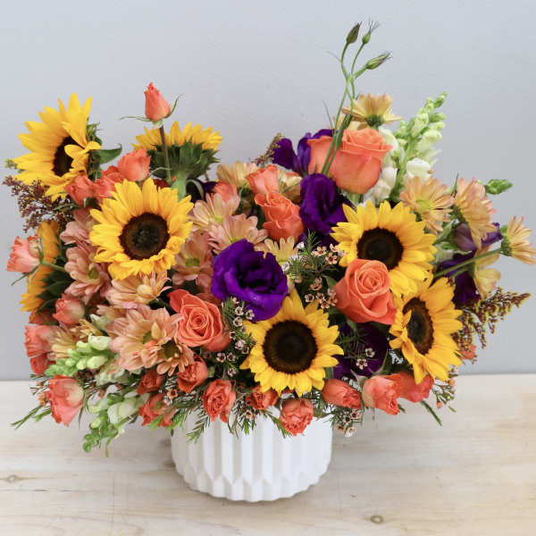 Bright arrangement of sunflowers, orange roses, and purple blooms in a white ceramic vase