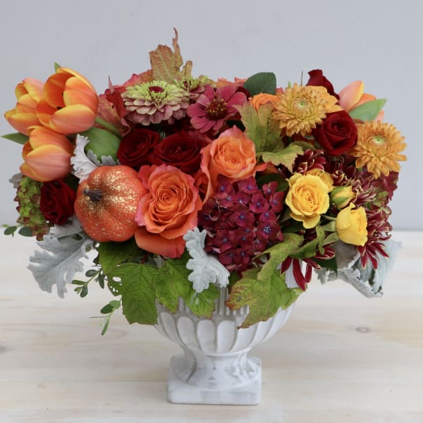 Low orange and red flower arrangement with roses and tulips in a white pedestal bowl.