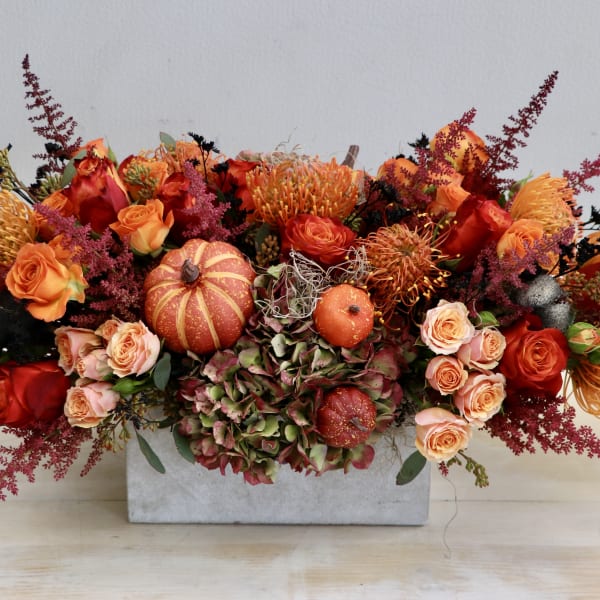 Low rectangular centerpiece with orange and red roses, protea, hydrangea, and small pumpkins.