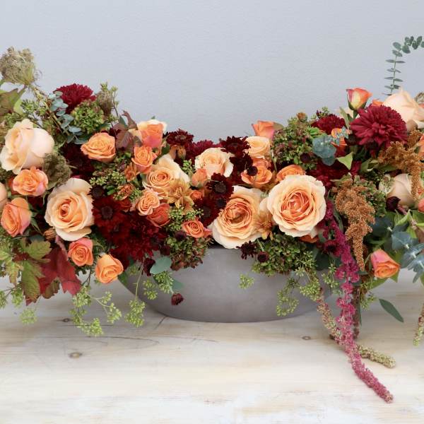 Low oval centerpiece of peach roses and burgundy flowers in a gray bowl-shaped container