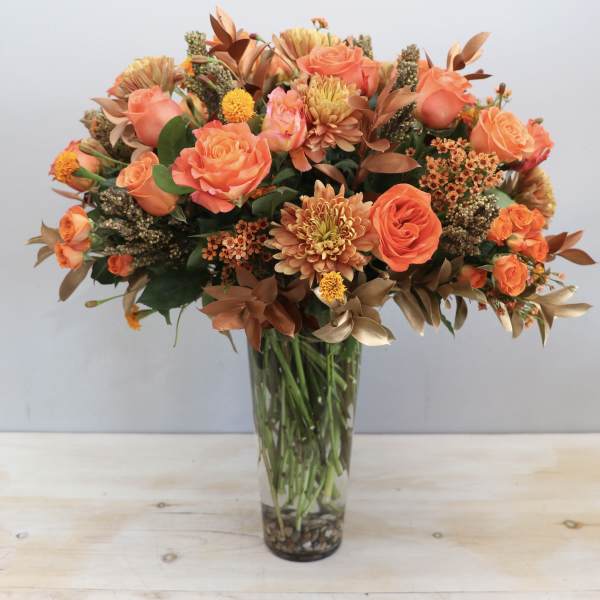 Tall vase arrangement of orange roses and chrysanthemums with bronze accents in a clear glass cylinder