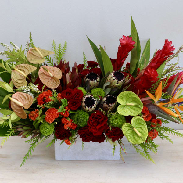 Long centerpiece of red and orange tropical flowers with green and peach anthuriums in a white rectangular vase