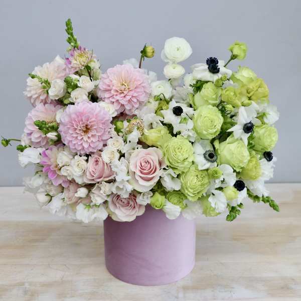 Round pastel arrangement of pink and green flowers in a pink hatbox-style container