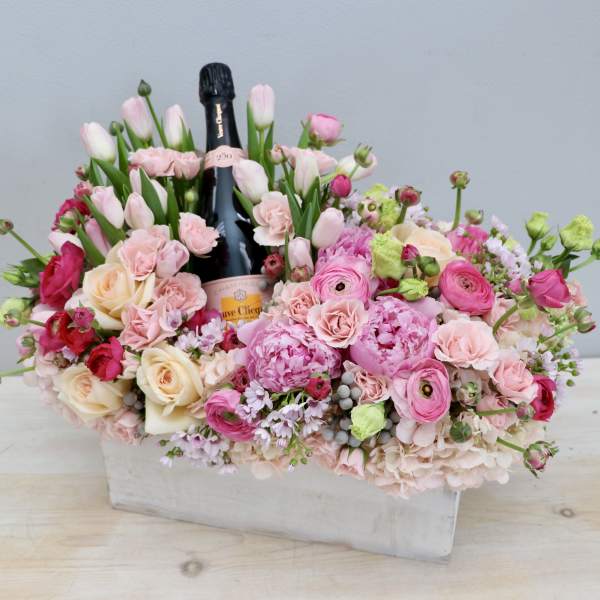 Pink and cream flower arrangement in a white box with a champagne bottle in the center