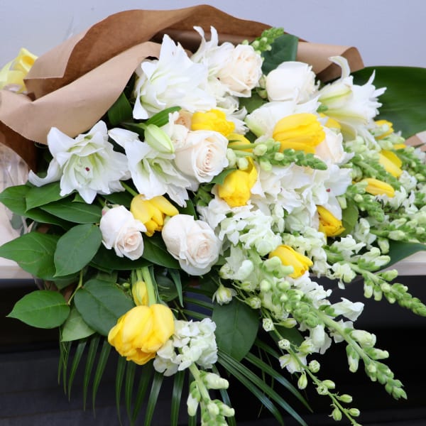 Wrapped bouquet of white lilies, cream roses, and yellow tulips with trailing white snapdragons