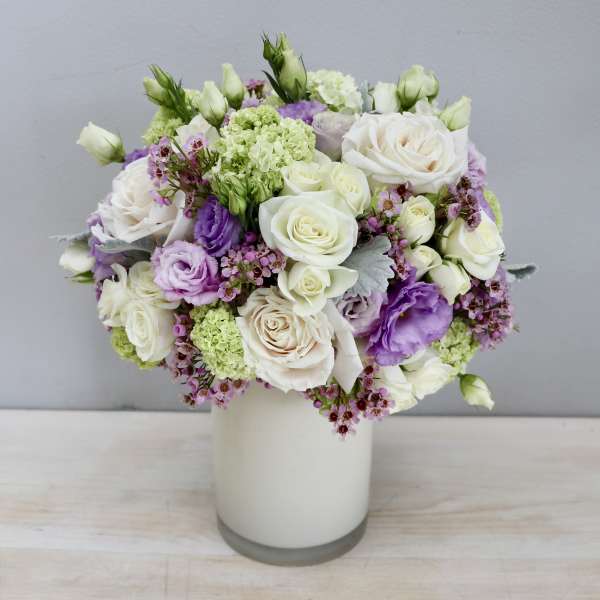 Compact arrangement of white roses and purple lisianthus in a white cylinder vase