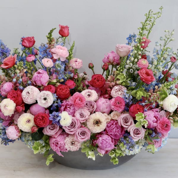 Low oval arrangement of pink and red roses, ranunculus, and peonies with blue delphinium in a gray bowl.