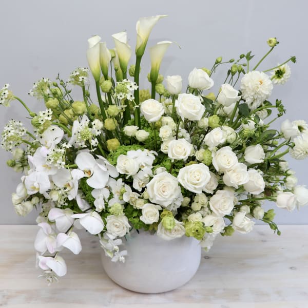 Large white and green arrangement of roses, calla lilies, orchids, and other blooms in a white ceramic bowl