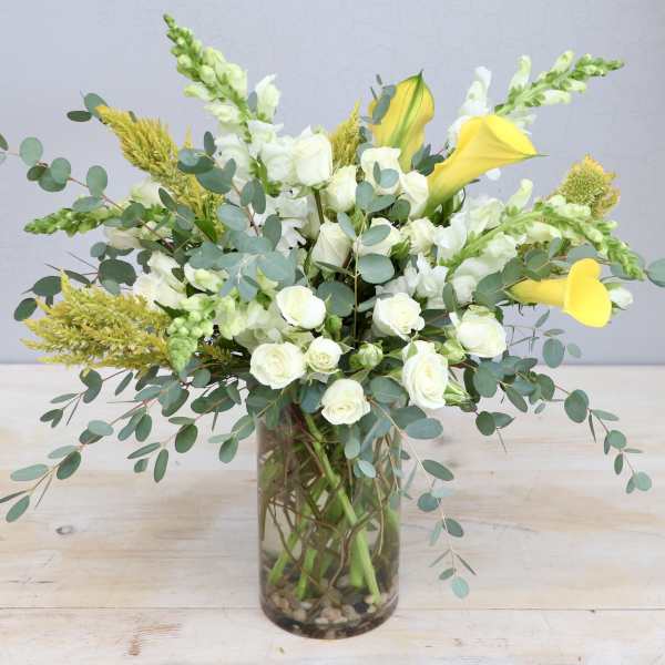Tall arrangement of white roses, white snapdragons, and yellow calla lilies in a clear glass vase