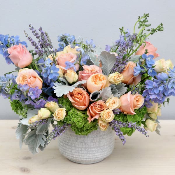 Low arrangement of peach roses and blue flowers in a white ceramic bowl vase