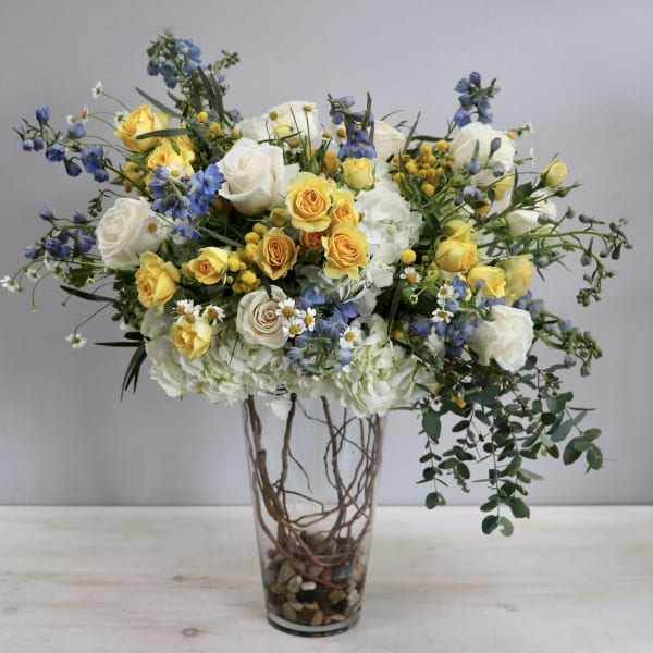 Tall arrangement of yellow and white roses, blue flowers, and hydrangeas in a clear glass vase.