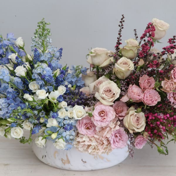 Low centerpiece with blue and white blooms on one side and pink and cream roses on the other in a white bowl