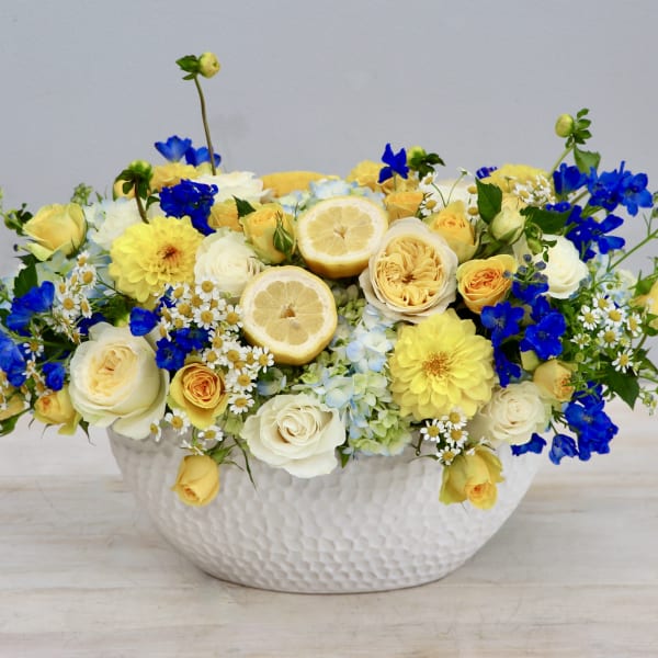 Low yellow, white, and blue flower arrangement with lemon slices in a white ceramic bowl