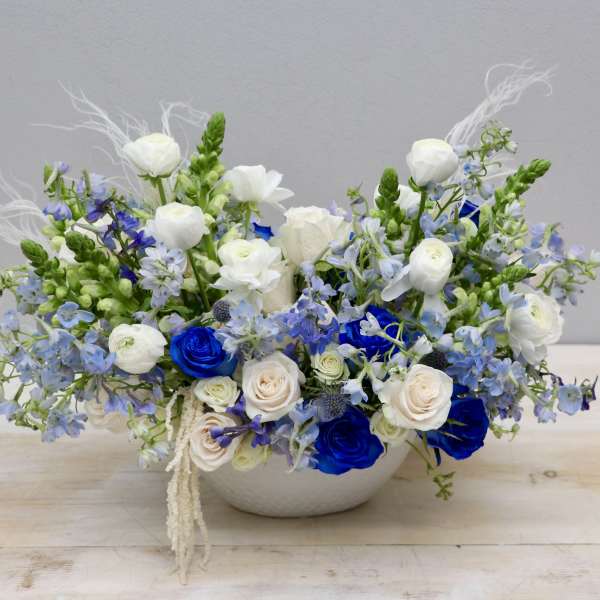 Low white bowl arrangement of blue and white roses with white ranunculus and light blue flowers