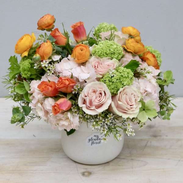 Low arrangement of peach roses, orange ranunculus, and green blooms in a white ceramic pot.