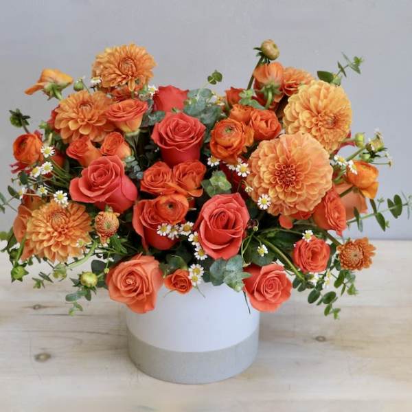 Orange roses, dahlias, and ranunculus in a low white cylinder vase with small white daisies