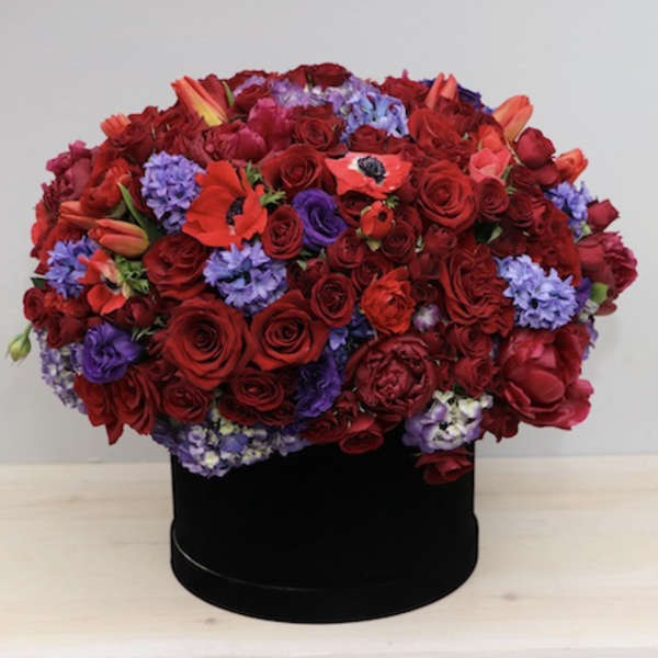 Low dome of red roses and purple blooms arranged in a black hatbox-style container
