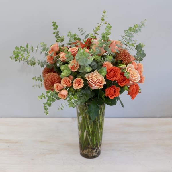 Tall glass vase arrangement of orange and peach roses with dahlias and eucalyptus.
