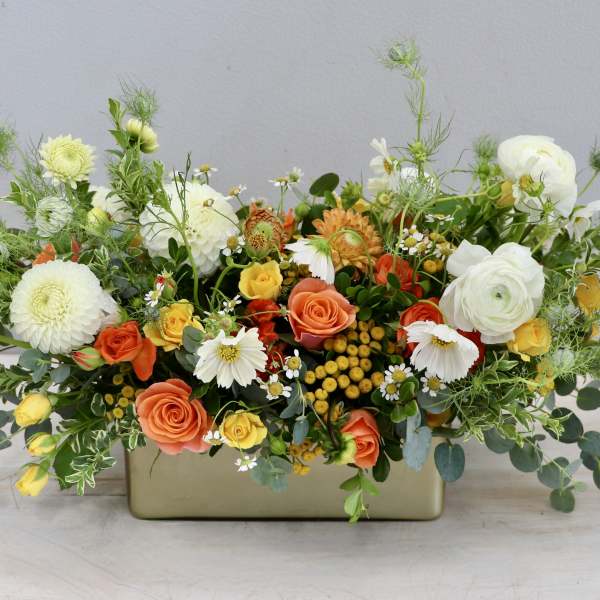 Low centerpiece of white dahlias, orange and yellow roses, and daisies in a gold rectangular vase