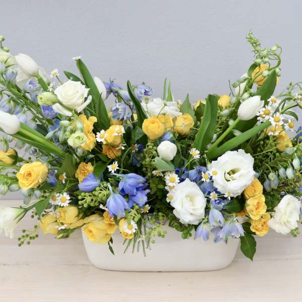 Low centerpiece of yellow roses, white tulips, and blue flowers in a white ceramic container