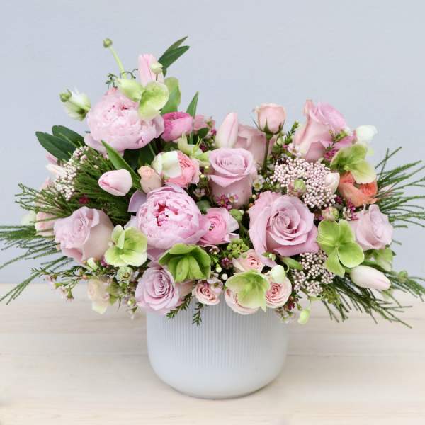 Low arrangement of soft pink roses, peonies, and tulips with light green blooms in a white ceramic pot
