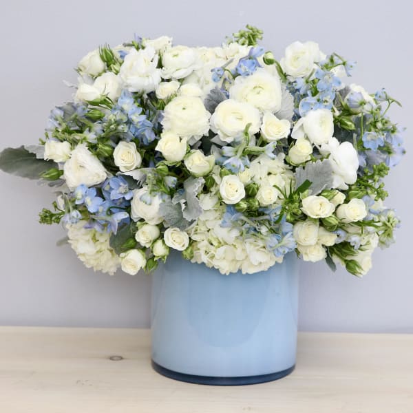 Round arrangement of white and light blue flowers in a light blue cylinder vase
