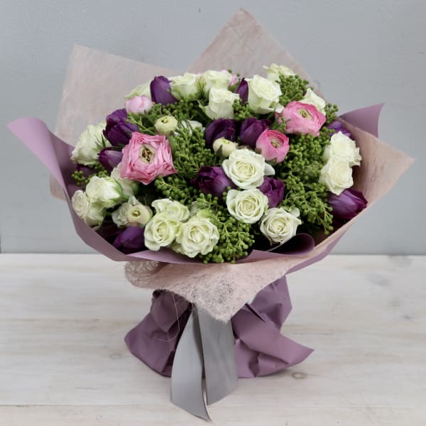 Hand-tied bouquet of white roses, purple tulips, pink ranunculus and green berries in mauve wrapping